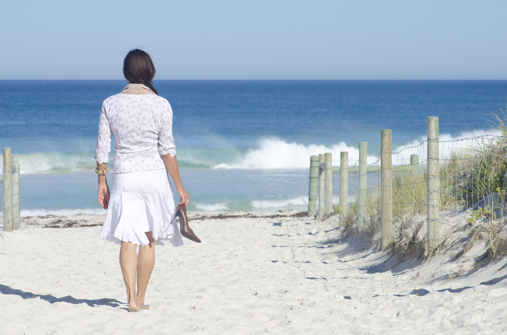Woman walking away towards beach barefoot, carrying her high heels